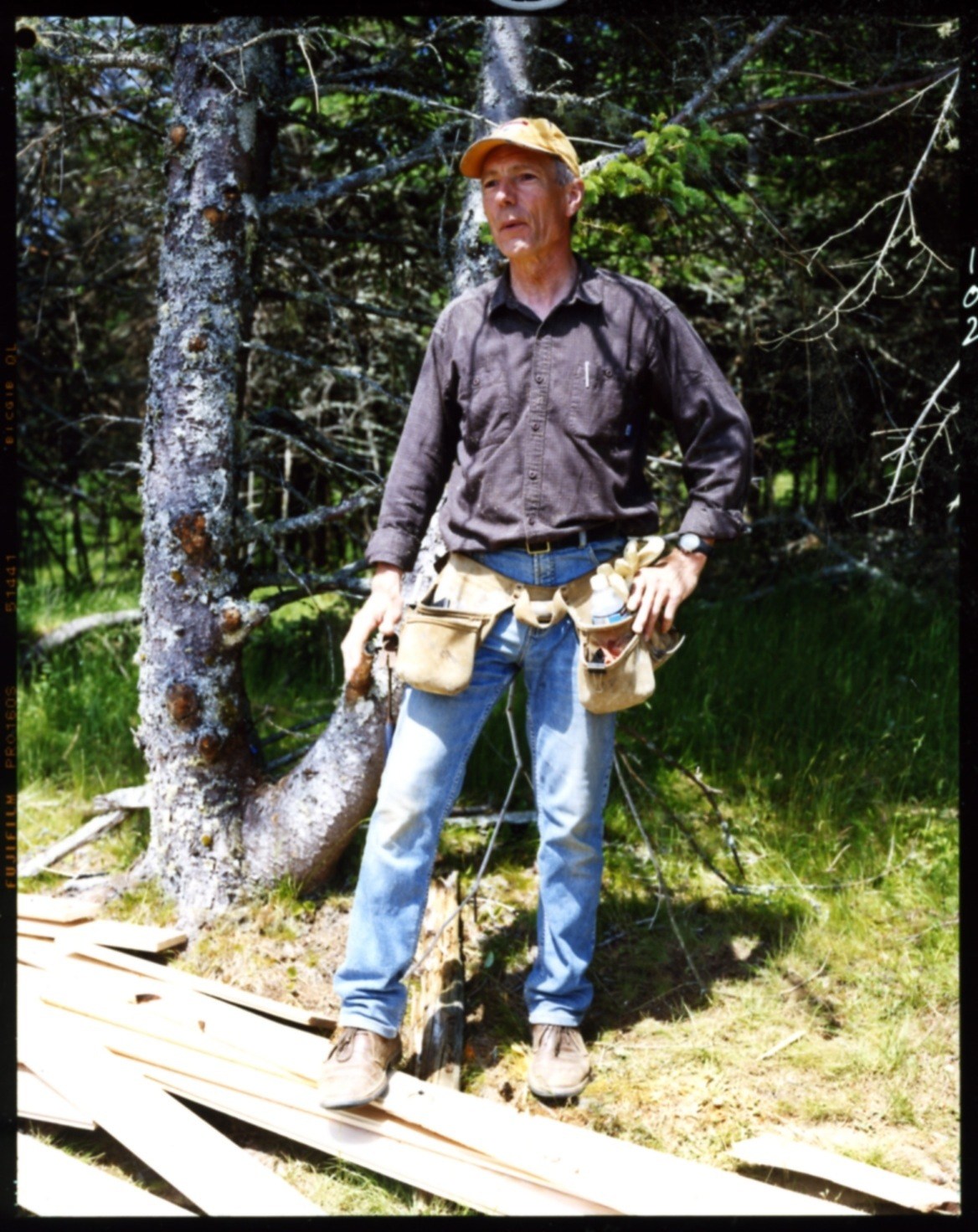 Builder Gordon MacLean wearing a tool belt and yellow hard hat stands in a forest setting.