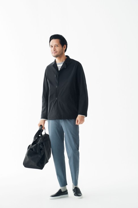 Man wearing black Teätora jacket, APC striped t-shirt, and Undecorated Man trousers holding a Tumi bag.