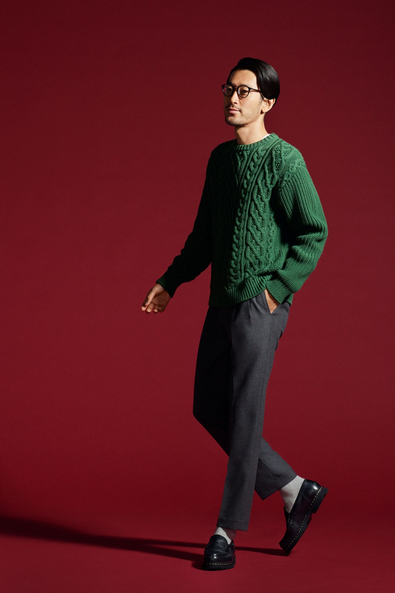Man in cream cable-knit sweater with dark trousers and brown shoes standing against red background