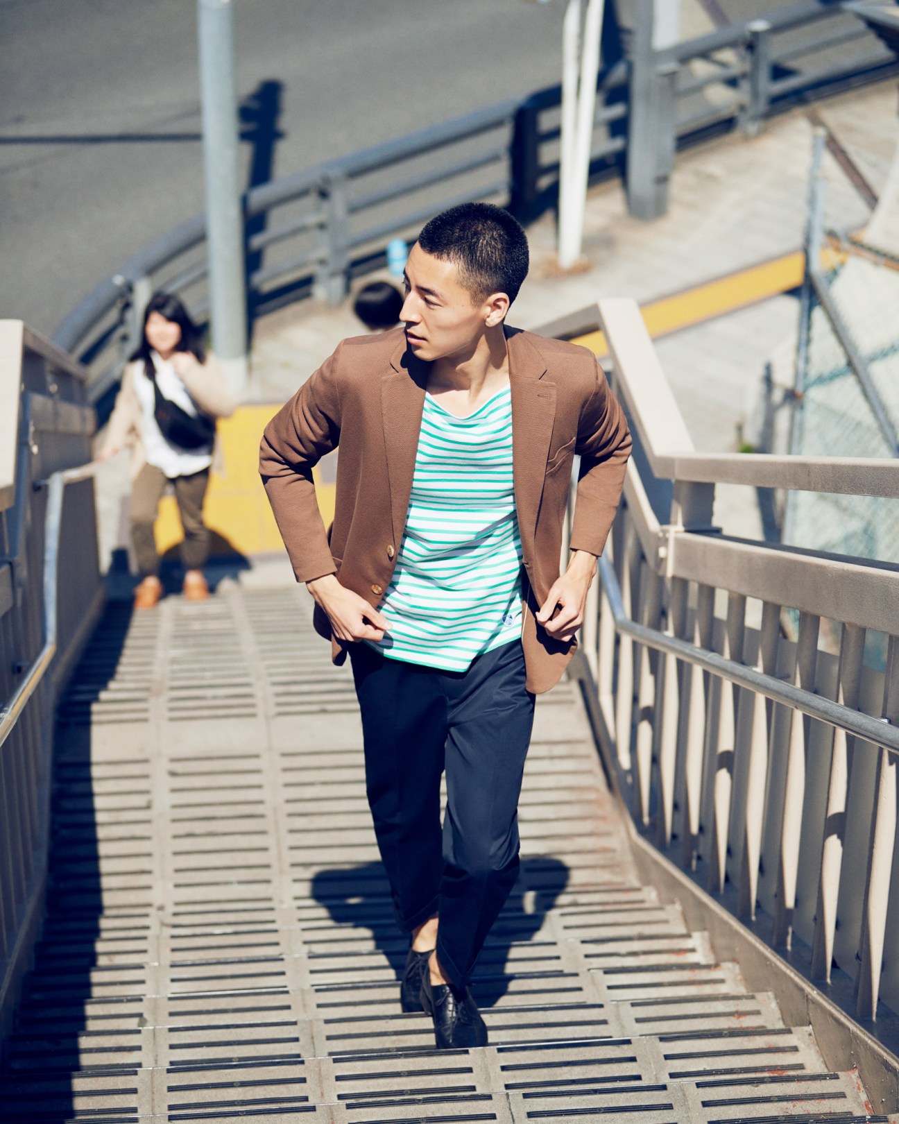 Man in brown jacket and striped t-shirt running on a steel pedestrian bridge in Tokyo.