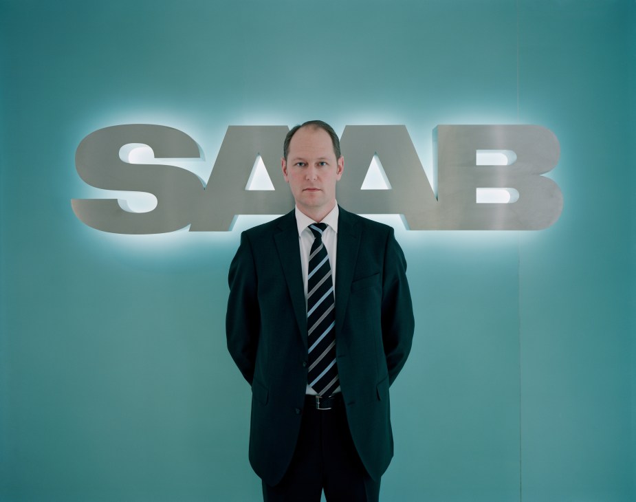 Man in business suit standing in front of illuminated SAAB logo against teal background.