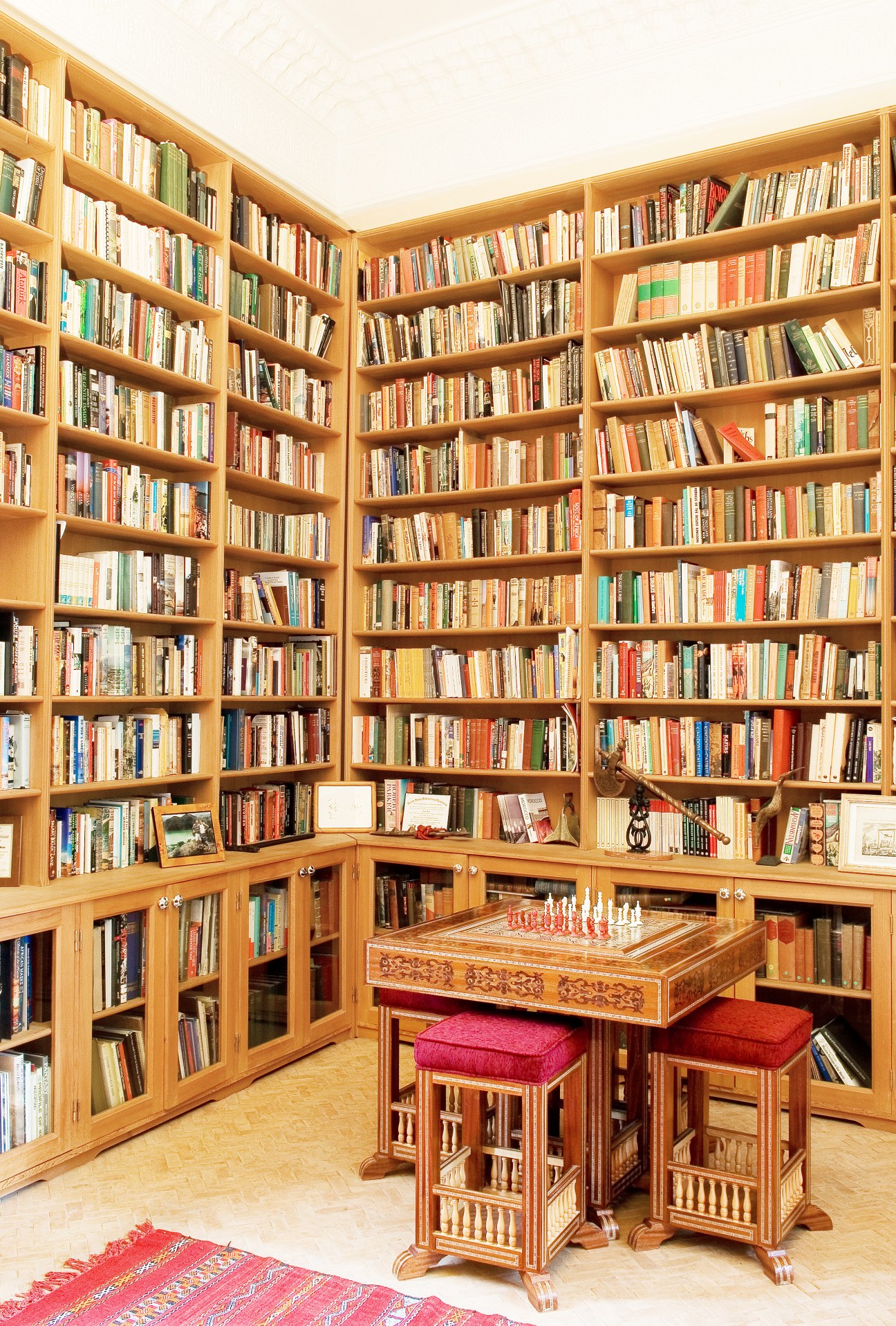 Wooden library shelves filled with books surrounding a central chess table with pink cushioned stools.