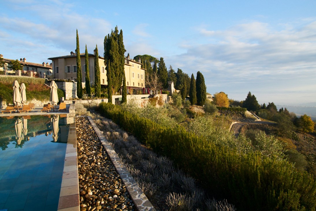 Tuscan villa with cypress trees, swimming pool, and terracotta buildings on hillside.