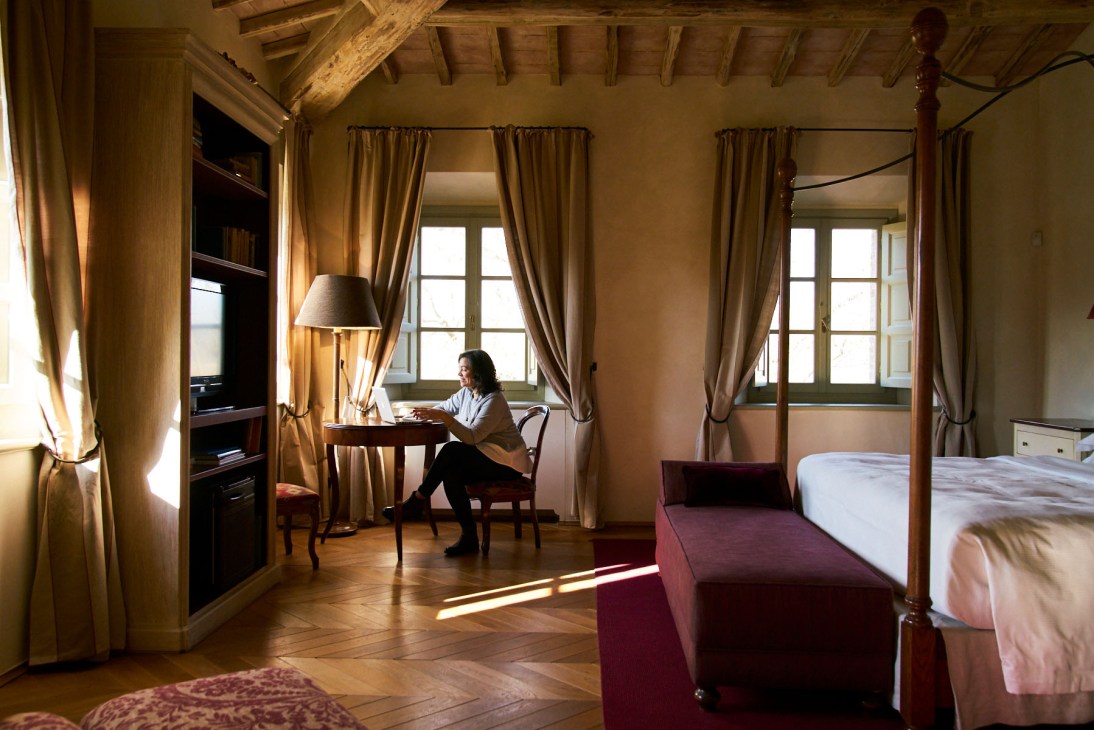 A woman sits at a desk inside a rustic Tuscan villa bedroom with wooden beams, cream curtains, and a four-poster bed.