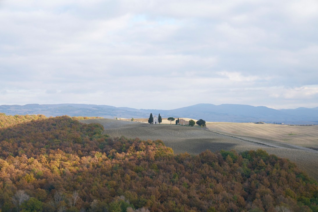 Rolling Tuscan countryside with autumn foliage, a solitary villa surrounded by cypress trees on a distant hilltop, and mountains on the horizon.