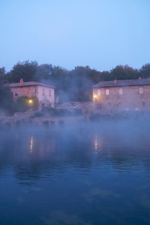 Stone buildings reflected in thermal pool at dawn with steam rising at Bagno Vignoni.