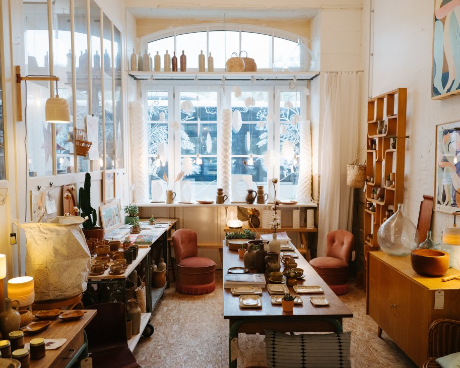 Interior of L'Atelier du Petit Parc boutique with wooden shelving, ceramics, and handcrafted items displayed near large windows.