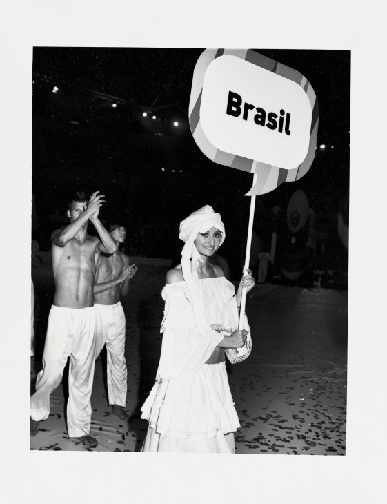 Woman in white dress and headwrap holds a speech bubble sign reading 'Brasil' while dancing at opening ceremony.
