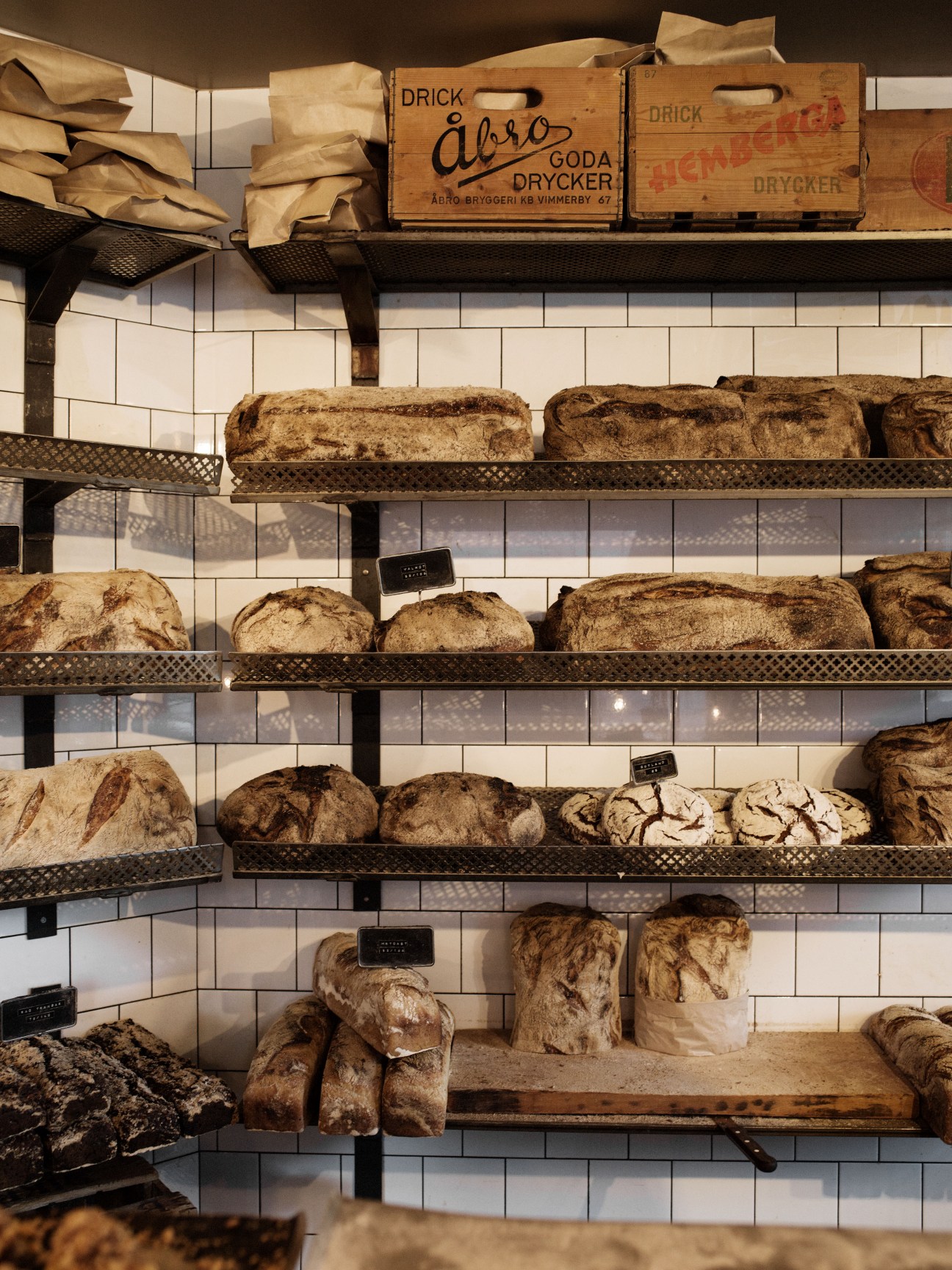 Rustic sourdough loaves arranged on metal shelves against white subway tiles with vintage wooden crates displayed above.