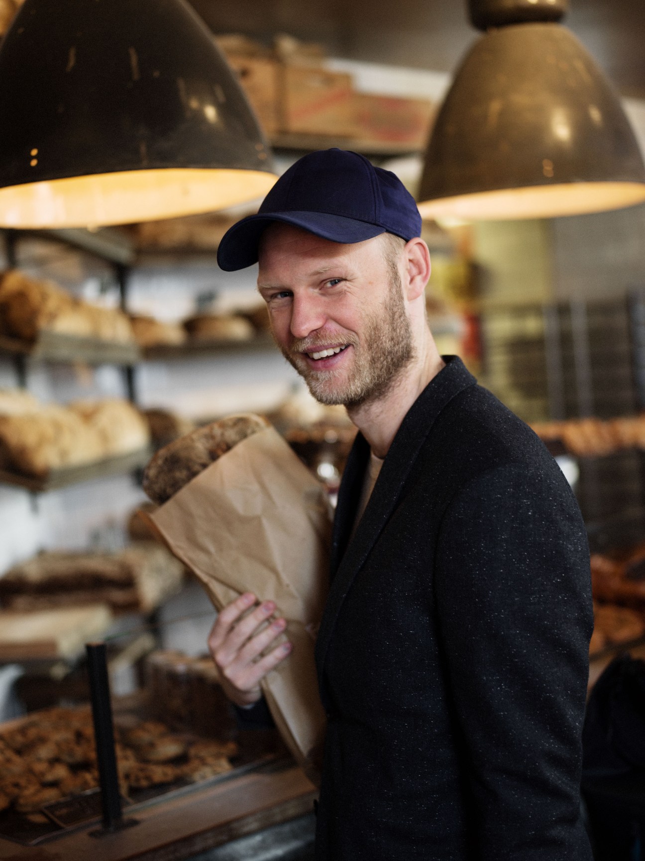 A smiling man wearing a navy cap and dark sweater holds a paper bag in a bakery with pastries and hanging lamps behind him.