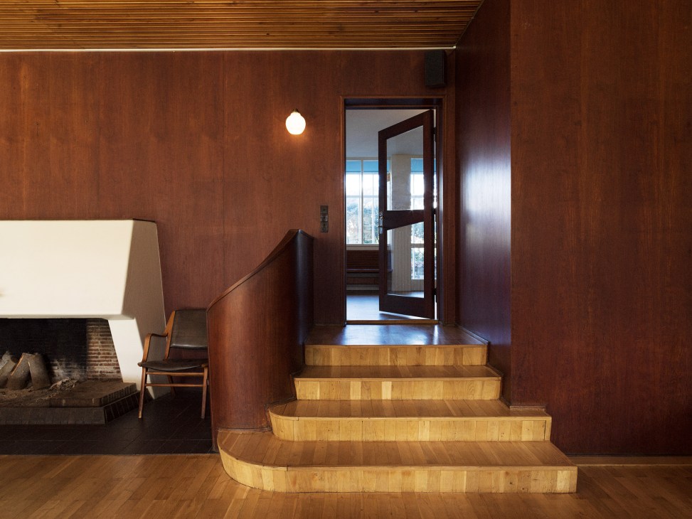 4th May College living room with wooden stairs, mahogany panelling, fireplace and modernist furniture