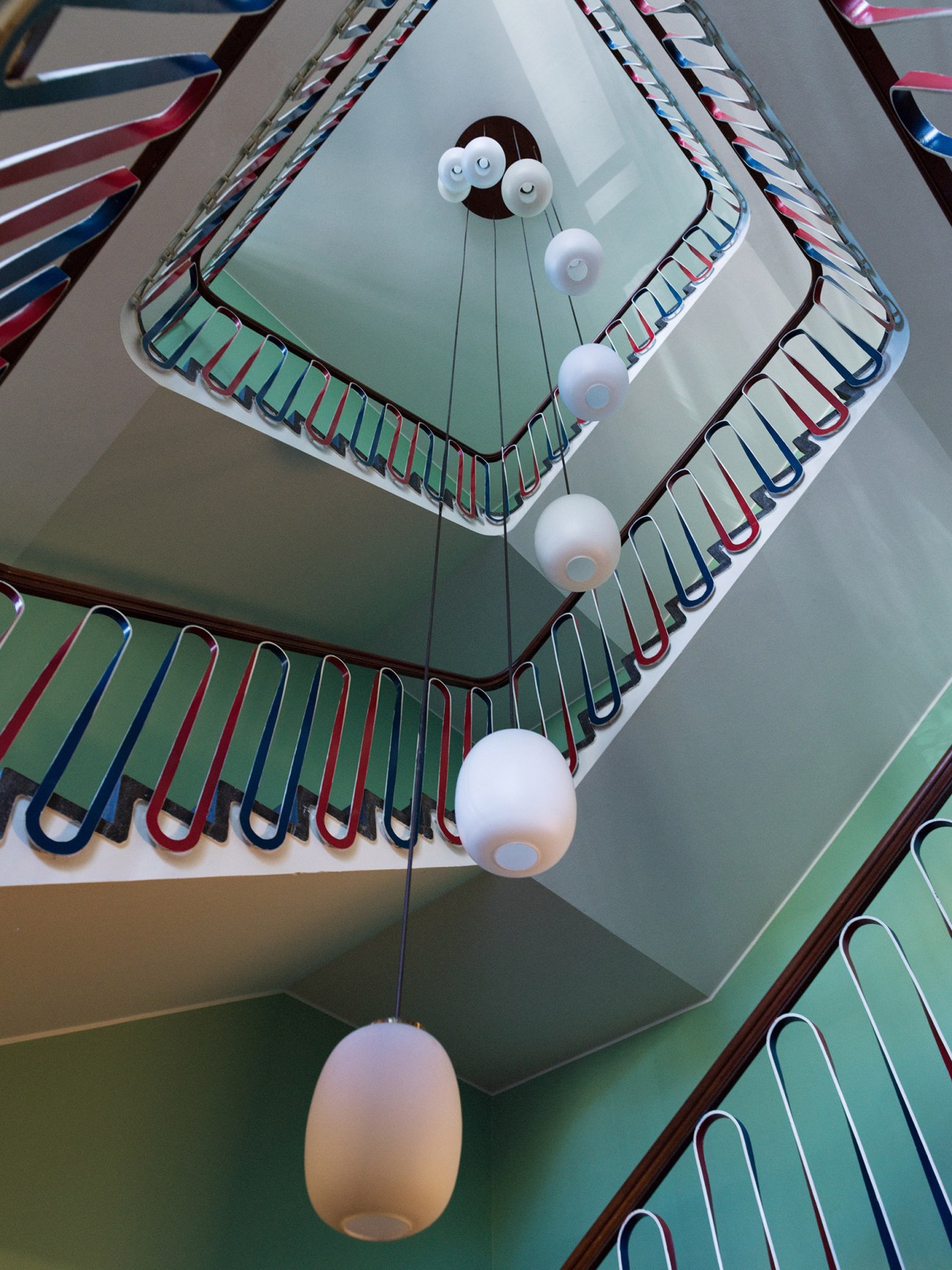 Overhead view of a spiral stairwell with mint green walls and red, white and blue railings, lit by white pendant lights.