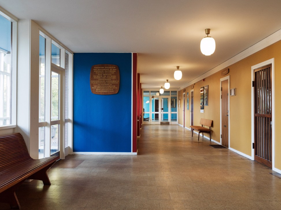 A hallway with blue and ochre walls, wooden doors, pendant lights, and a memorial plaque in 4th May College, Copenhagen.