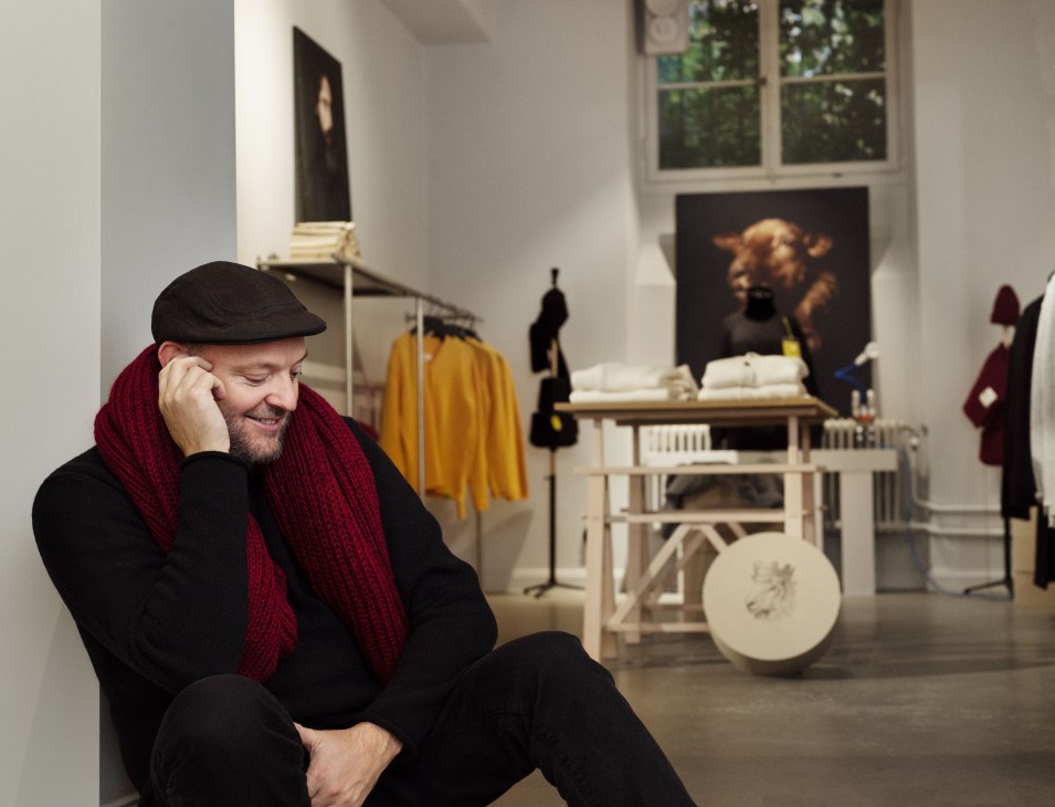 Alexander Stutterheim sits in the John Sterner shop wearing a red scarf, surrounded by displayed knitwear and clothing racks.