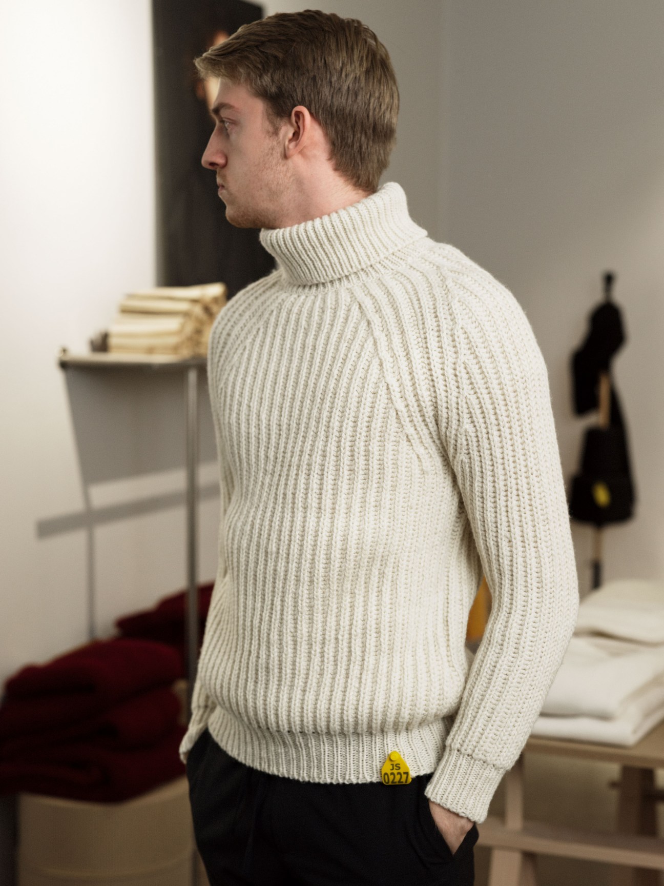 Man wearing a cream ribbed turtleneck sweater with a yellow sheep tag label at the hem, shown from behind.
