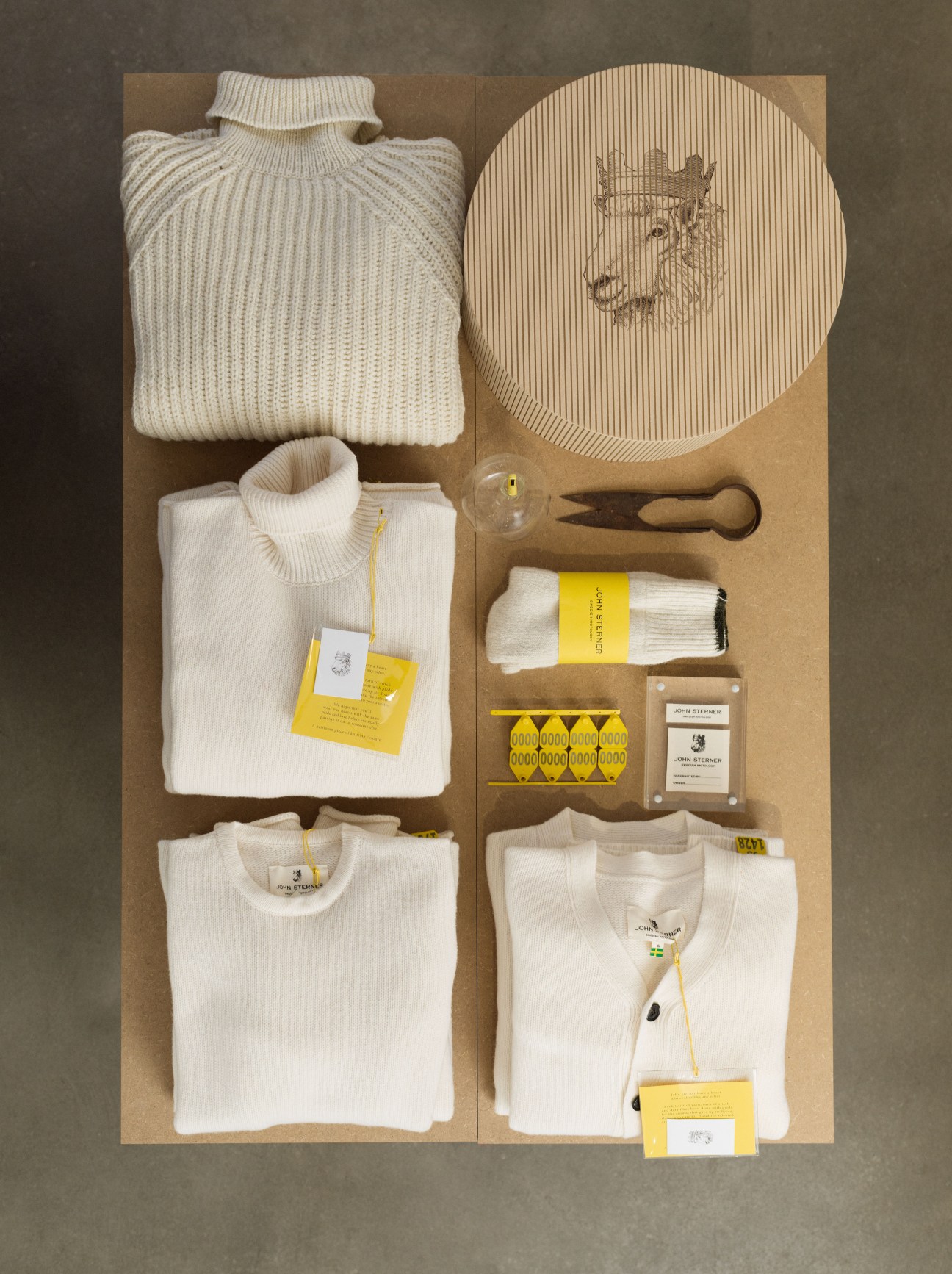 John Sterner knitwear collection displayed flat on kraft paper, including cream sweaters, socks, yarn, circular knitting pattern, wooden tools, and yellow branded tags.