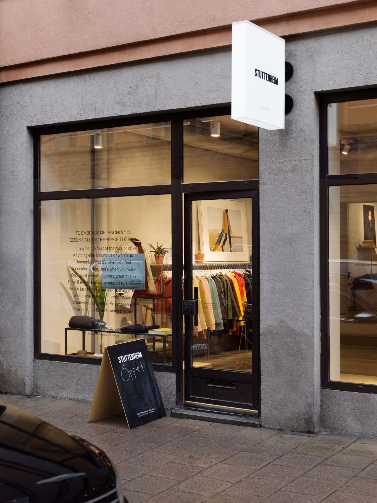 Exterior storefront of Stutterheim/John Sterner shop with large windows displaying knitwear and raincoats on a Stockholm street.