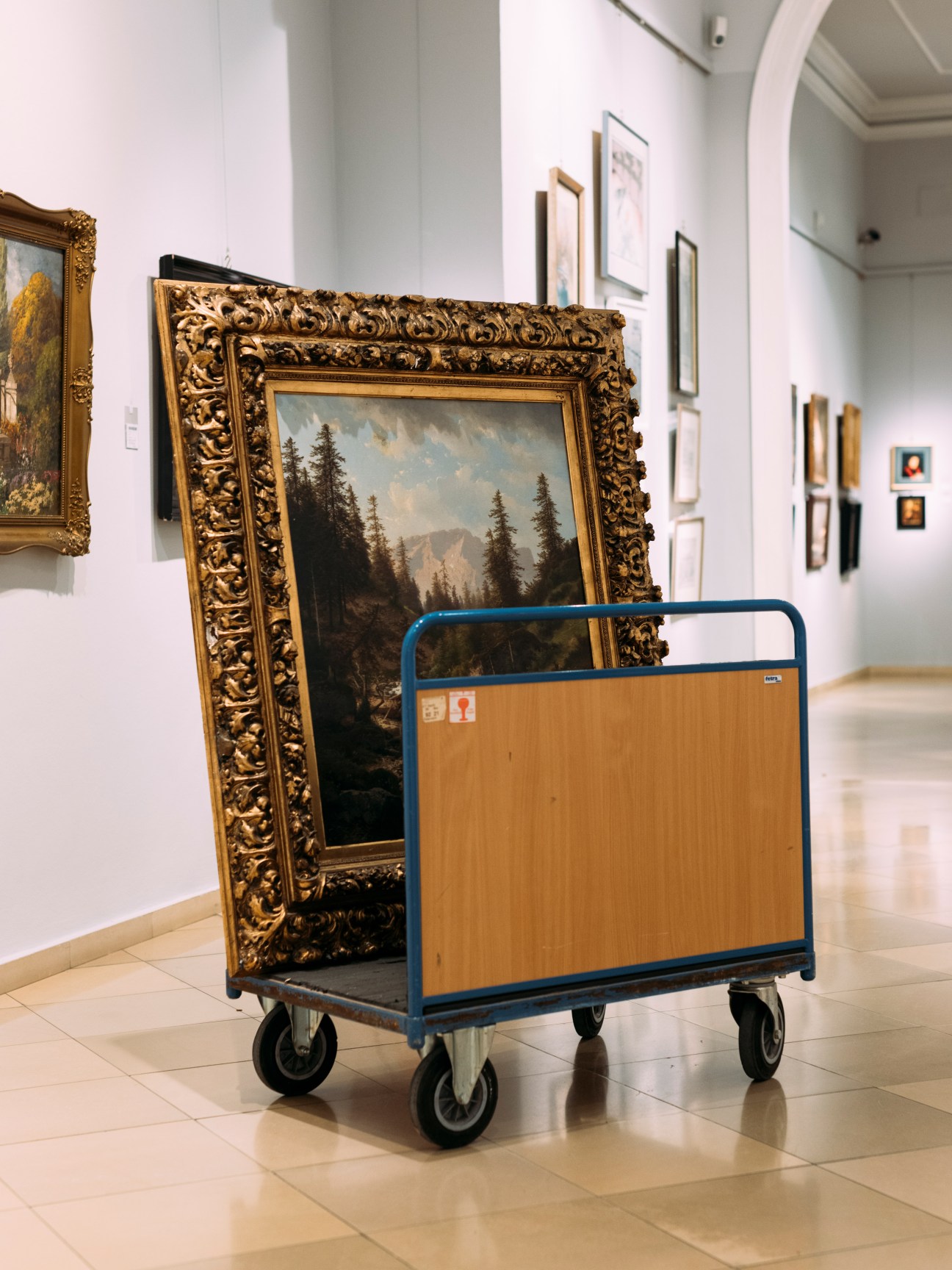 Ornately framed landscape painting on wheeled cart in white-walled museum gallery with other artworks