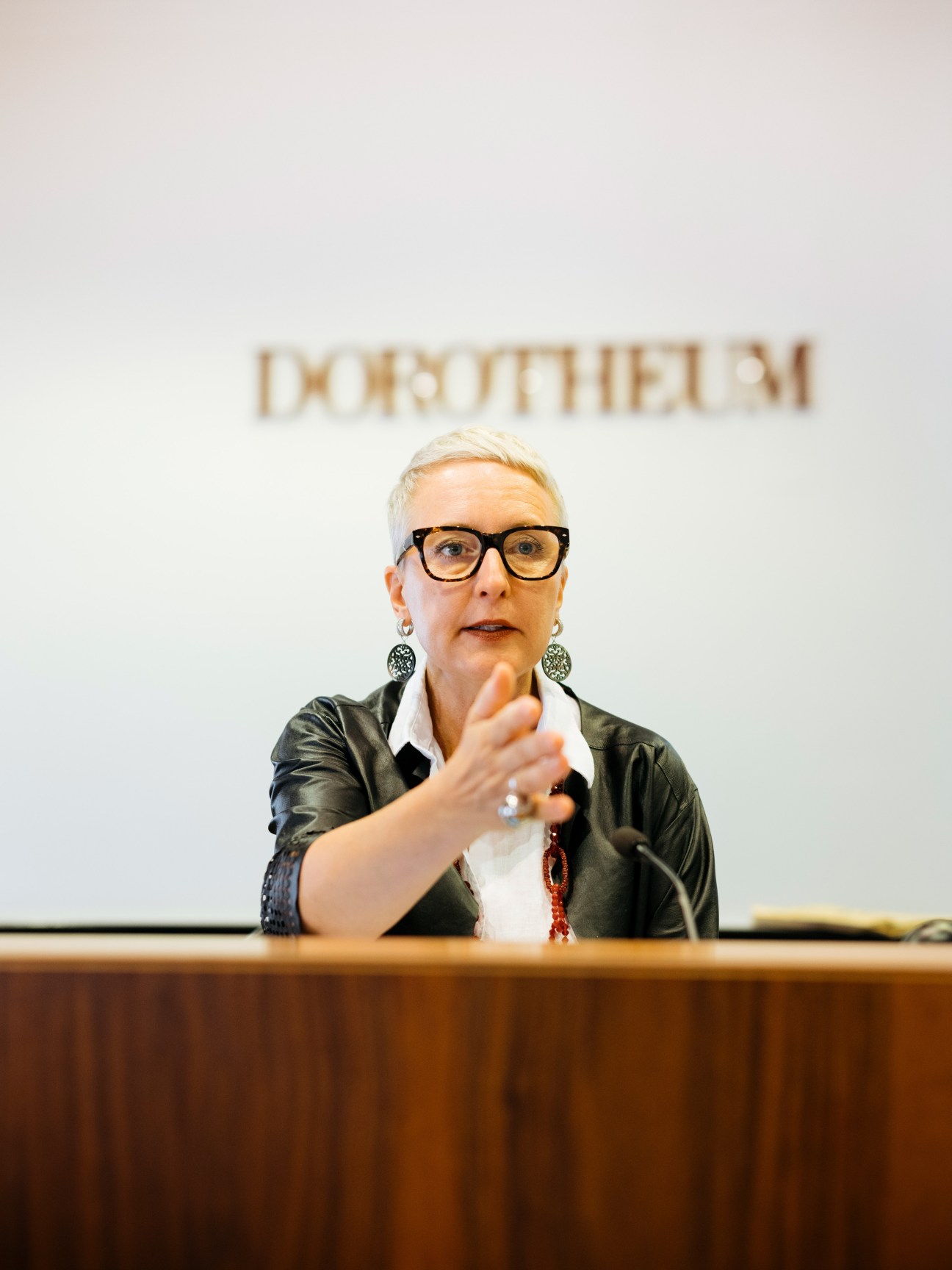 Woman with blonde hair and glasses speaking at auction podium with hand gesture