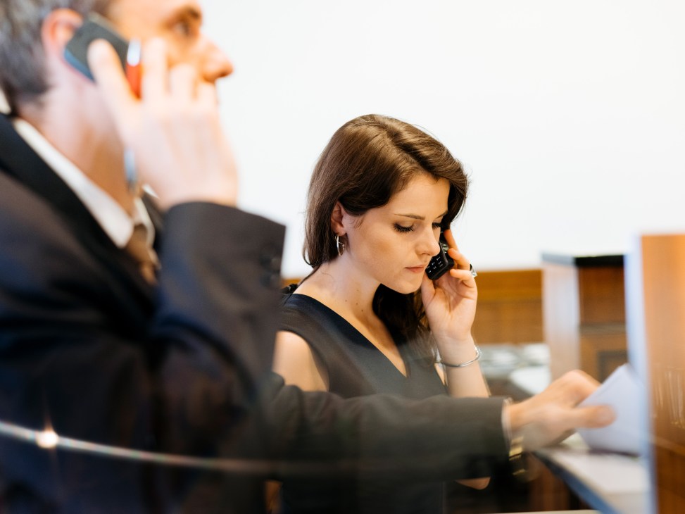 Woman in black dress taking phone bids at auction house with man in suit also on phone in background