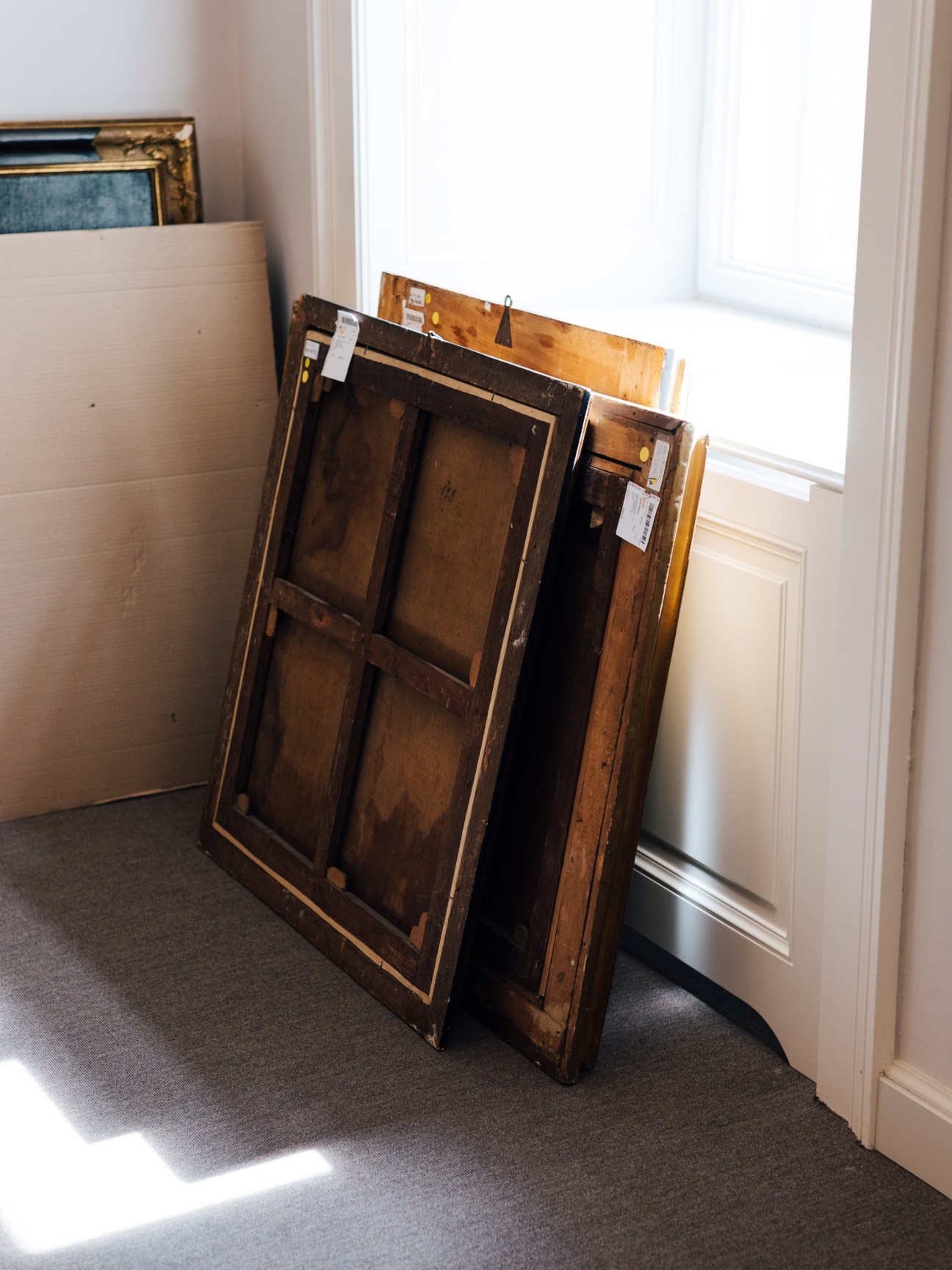 Framed paintings leaning against a white wall near a window in an auction house storage area