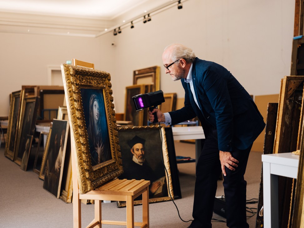 Mark MacDonnell examining a painting with UV light in a room filled with artwork and frames