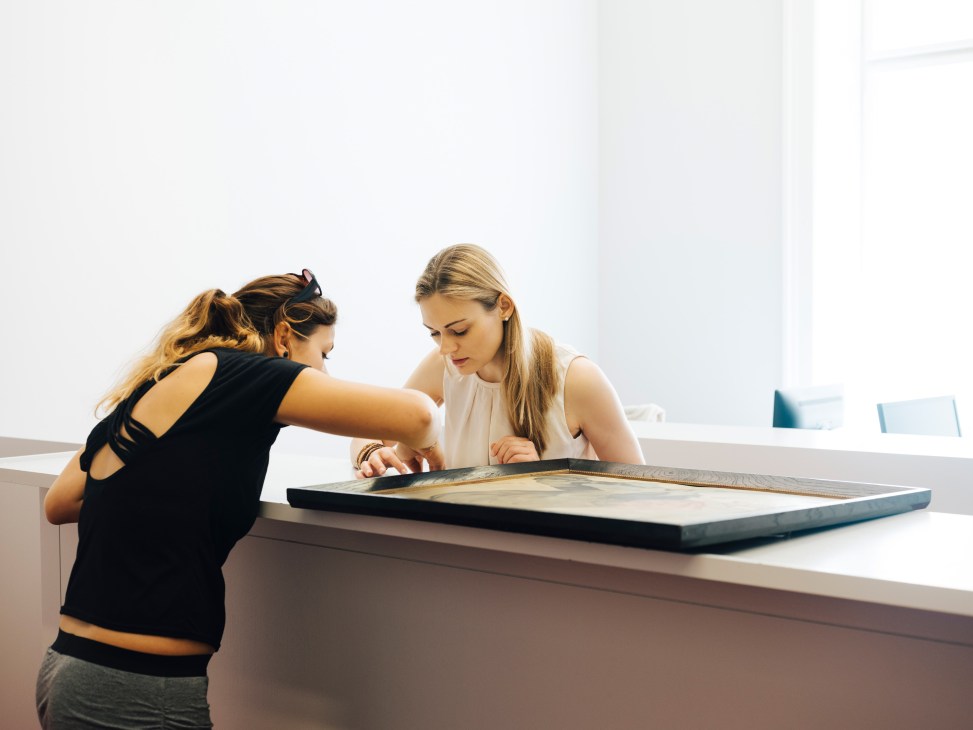 Two women examining a framed painting on a white surface in a gallery setting
