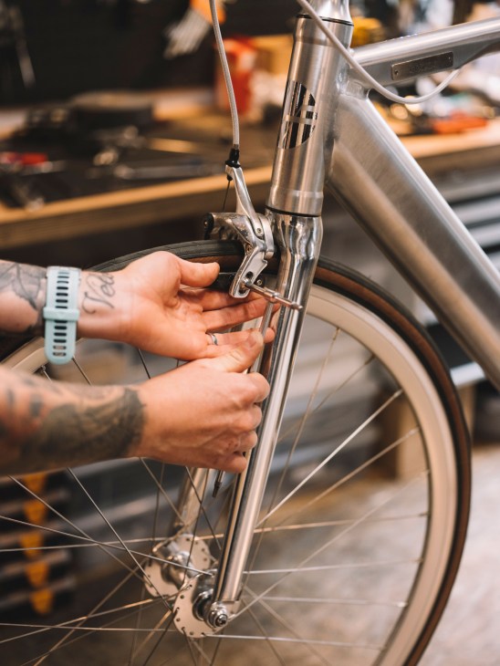 A tattooed mechanic's hands adjusting the brakes on a sleek silver e-bike frame.