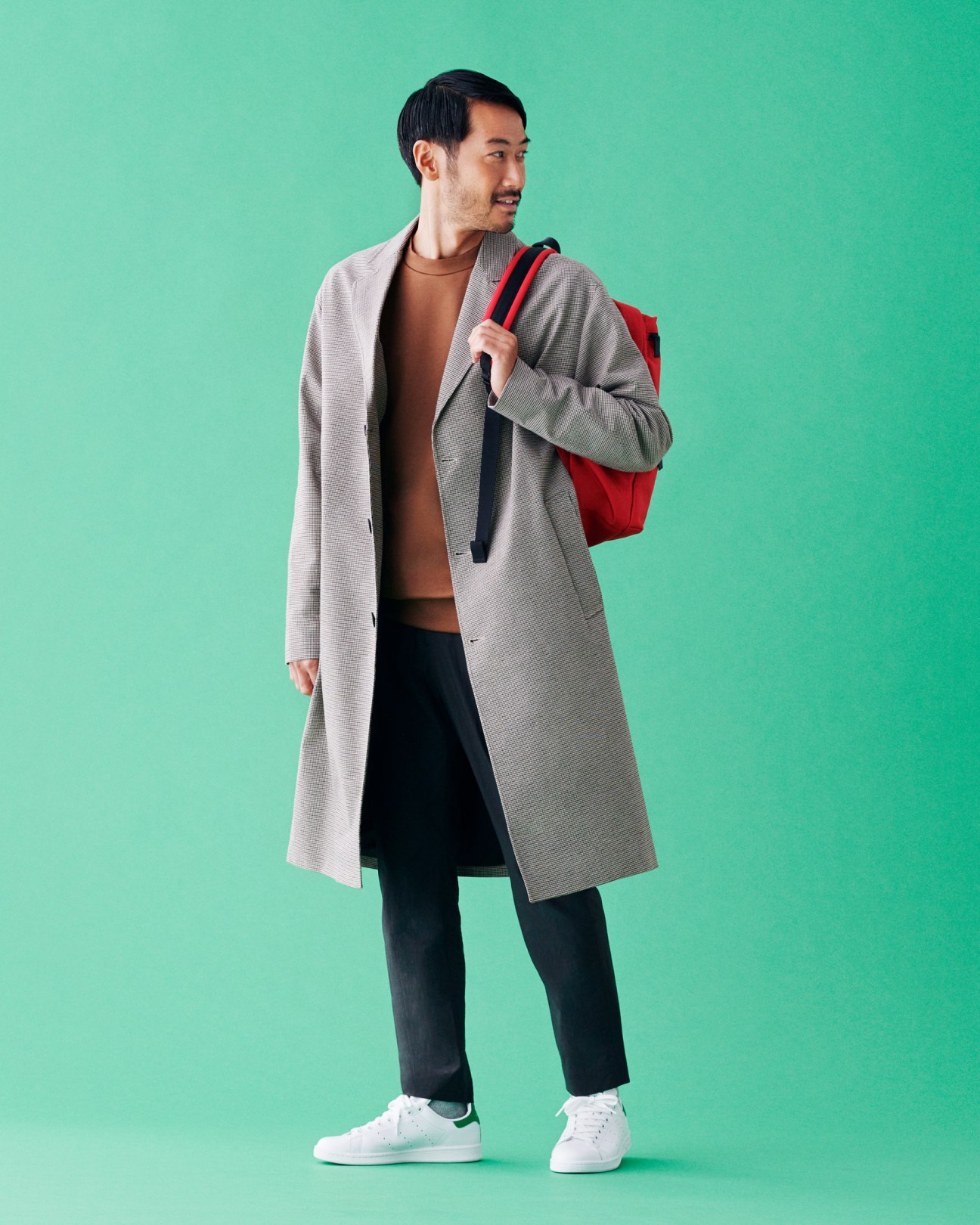 Man wearing gray coat, brown t-shirt, black trousers, white sneakers and red backpack against mint green background.