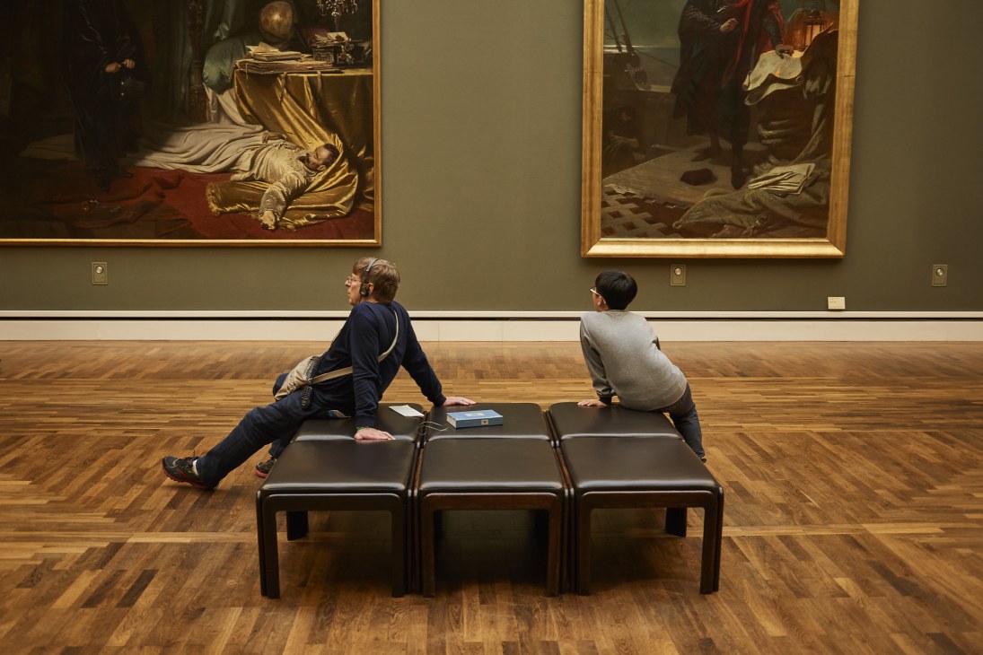 Two visitors viewing classical paintings in a museum gallery with wooden flooring.