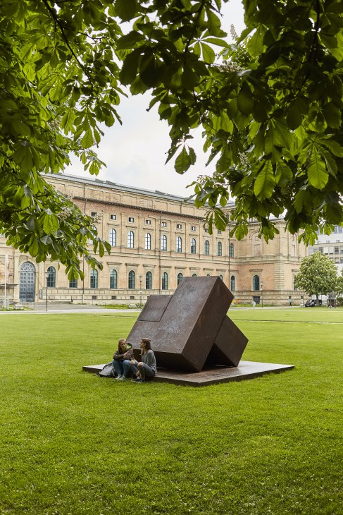 Two people sit beneath a large dark metal sculpture on a lawn with a classical stone building behind