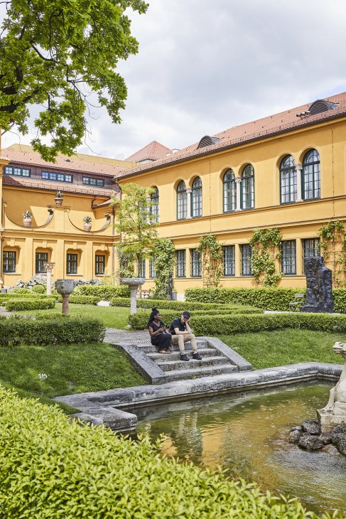 Two people sit by a pond in the manicured courtyard garden of Lenbachhaus, a yellow Tuscan-style villa in Munich.