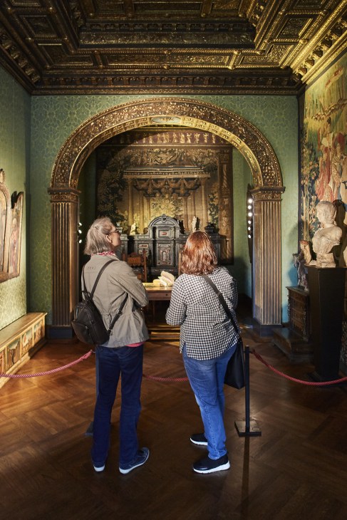 Two visitors viewing ornate room with gilded arch and classical sculptures at Lenbachhaus museum.
