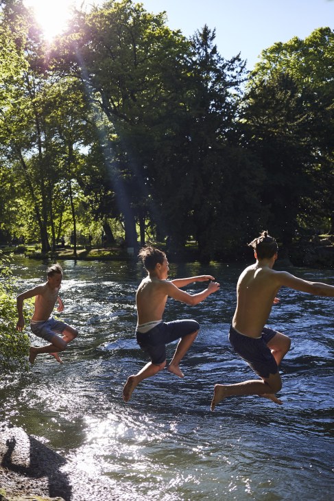 Three shirtless boys running into a river surrounded by lush green trees on a sunny day.