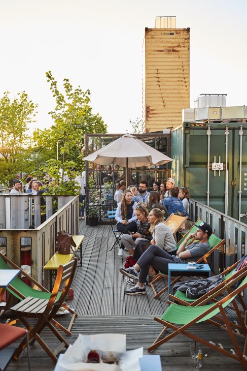 People gather on a rooftop container venue with deck chairs, umbrellas and greenery.