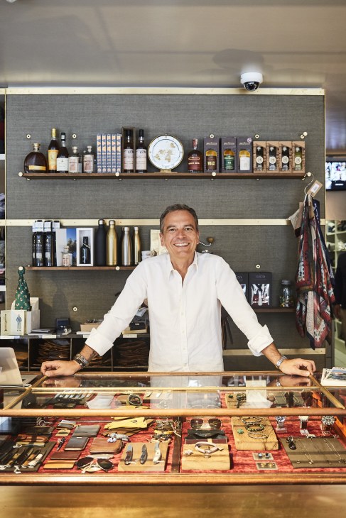 Man in white shirt standing behind retail counter with shelves of products and display items behind him.