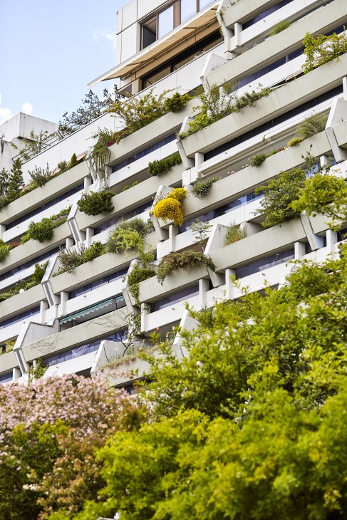 Modern residential building with tiered balconies featuring abundant green plants and flowers.