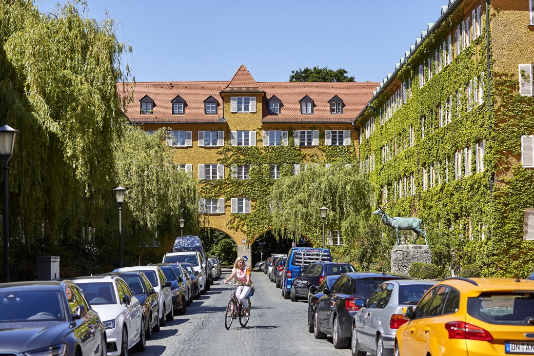 A tree-lined courtyard with ivy-covered historic buildings, parked cars, and a cyclist riding through the center.
