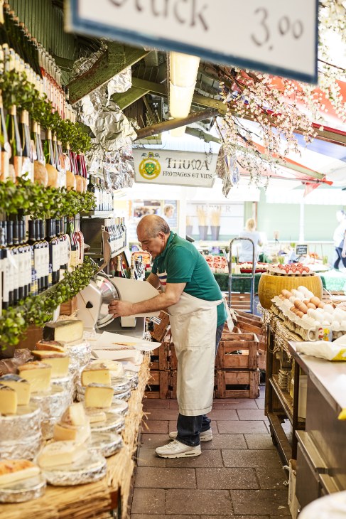 Man in green shirt and apron slicing cheese at a market stall with hanging herbs and produce displays.
