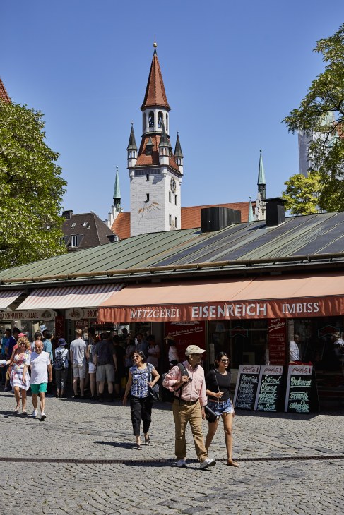 Munich's Viktualienmarkt food market with historic church tower and visitors shopping at stalls.