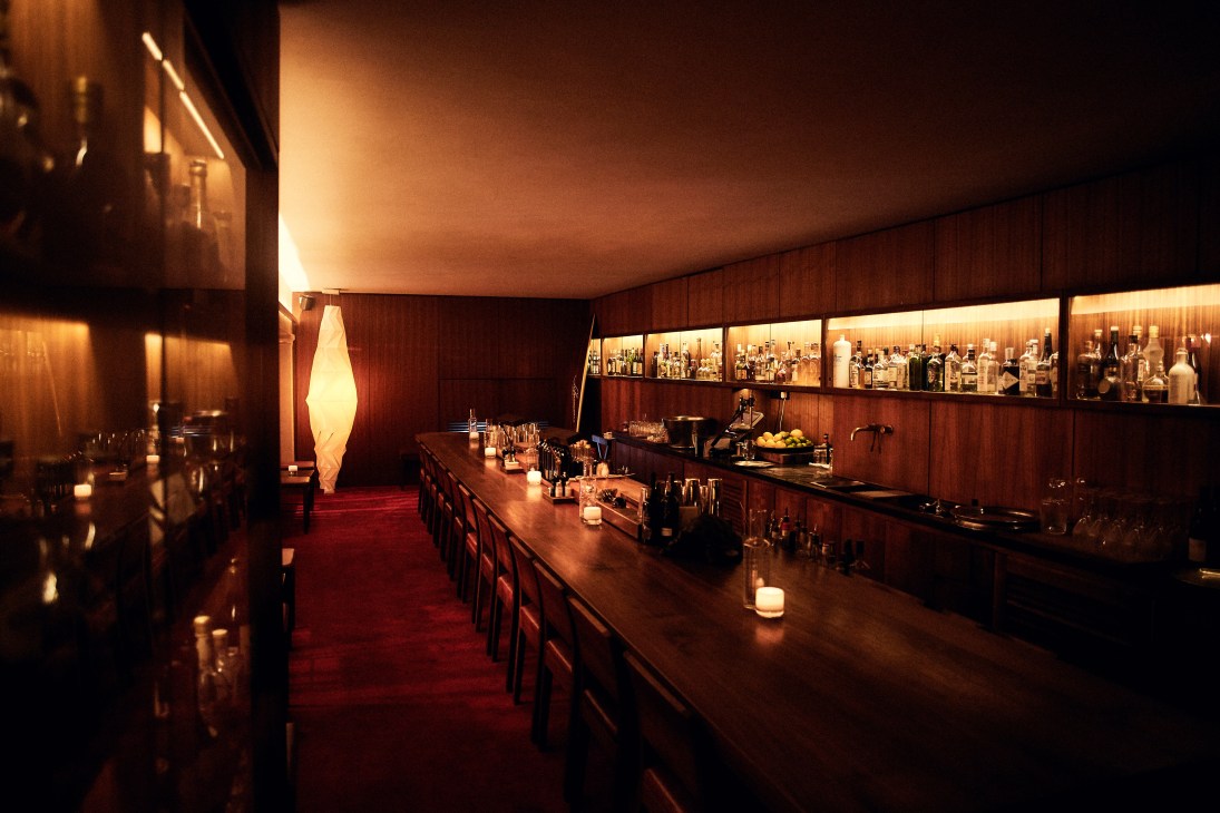 Dimly lit bar interior with wooden walls, bar counter, bottles on shelves, and warm ambient lighting