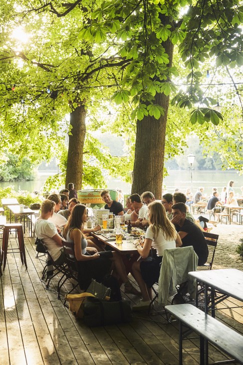 People dining outdoors under large green trees at Seehaus beer garden with water visible in background.