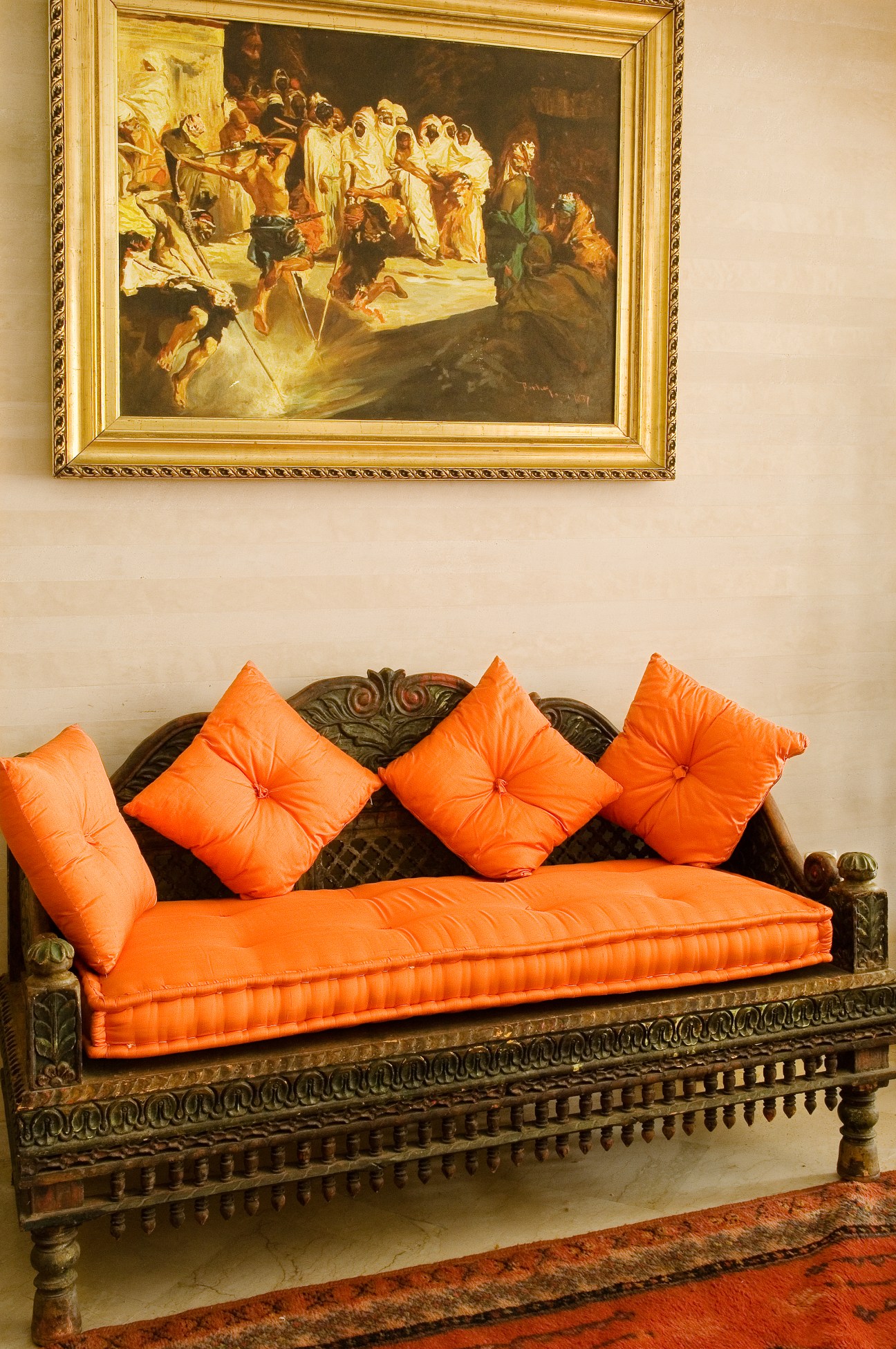 Ornate wooden daybed with bright orange cushions beneath a framed classical painting on a cream wall.