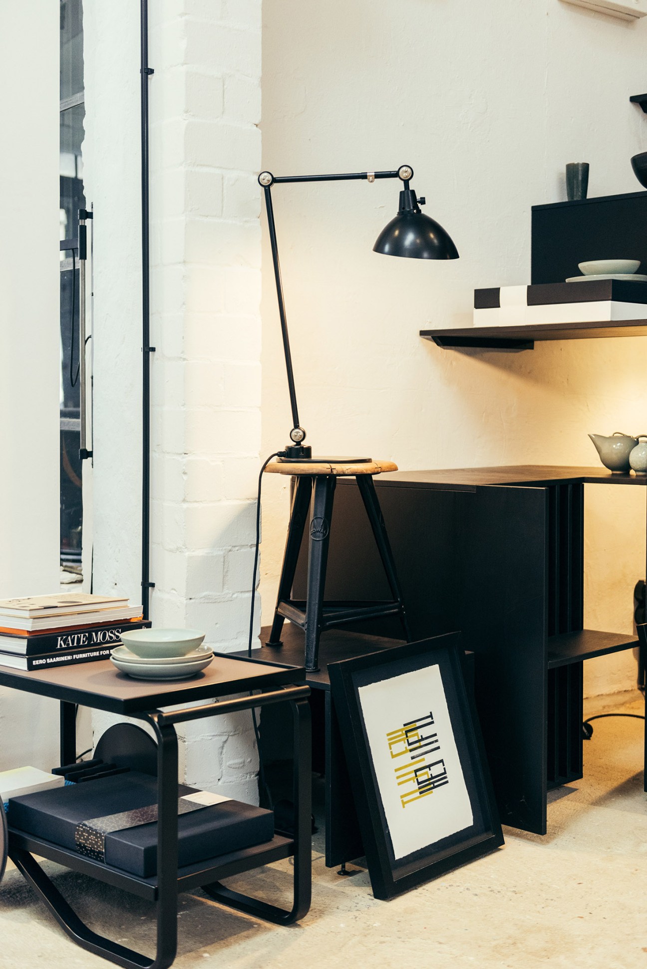 A black articulated Midgard lamp positioned above a desk with books and dishware in a showroom setting.