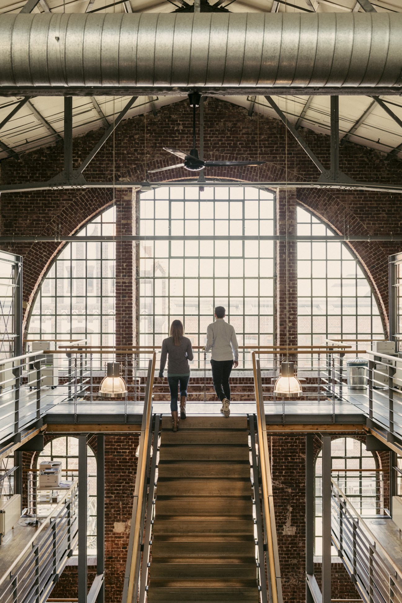 Two people jogging up wooden stairs inside a converted industrial building with large arched windows and exposed brick walls.