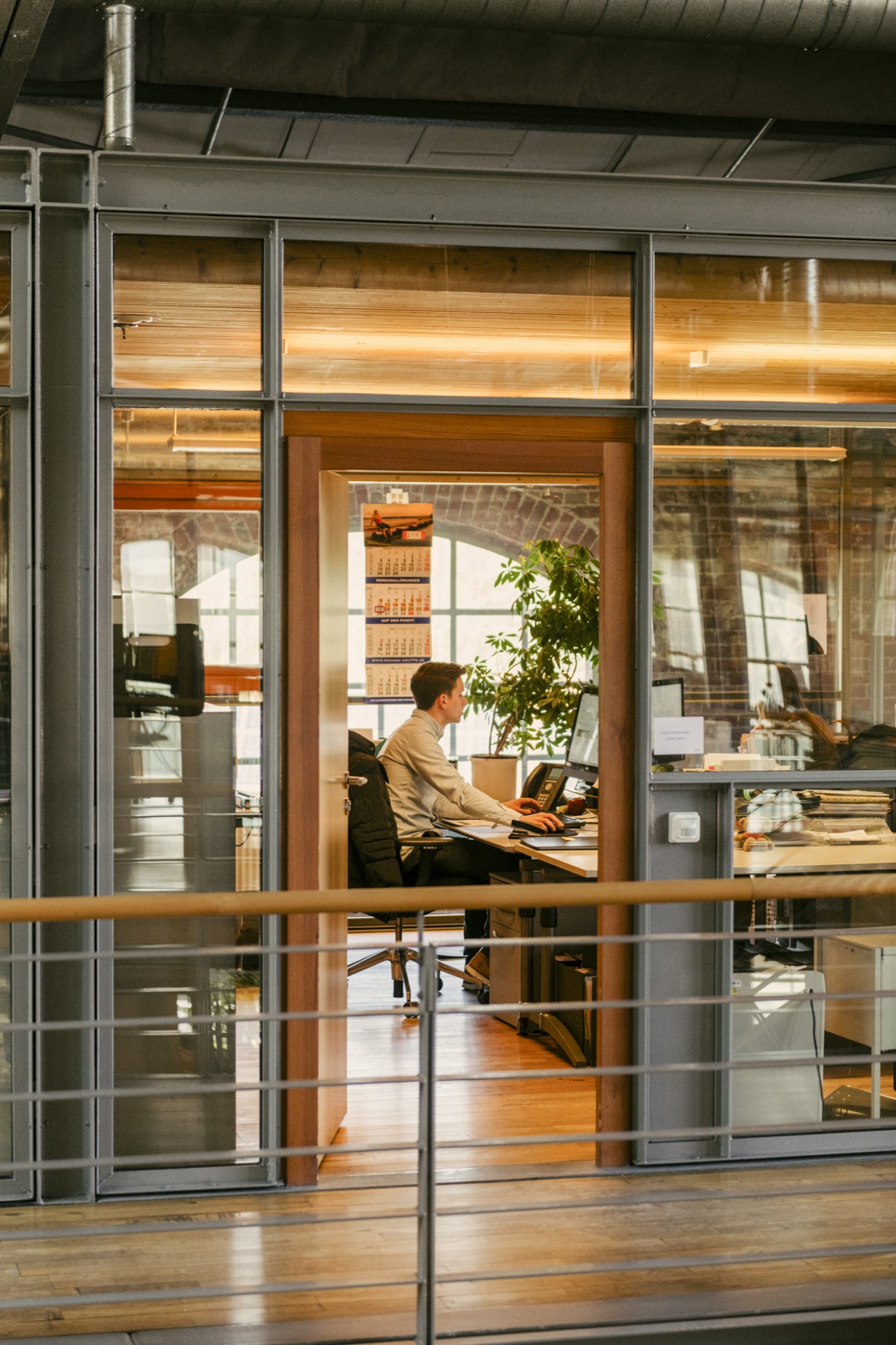 Man working at a desk in a glass-walled office cube inside a converted industrial building.