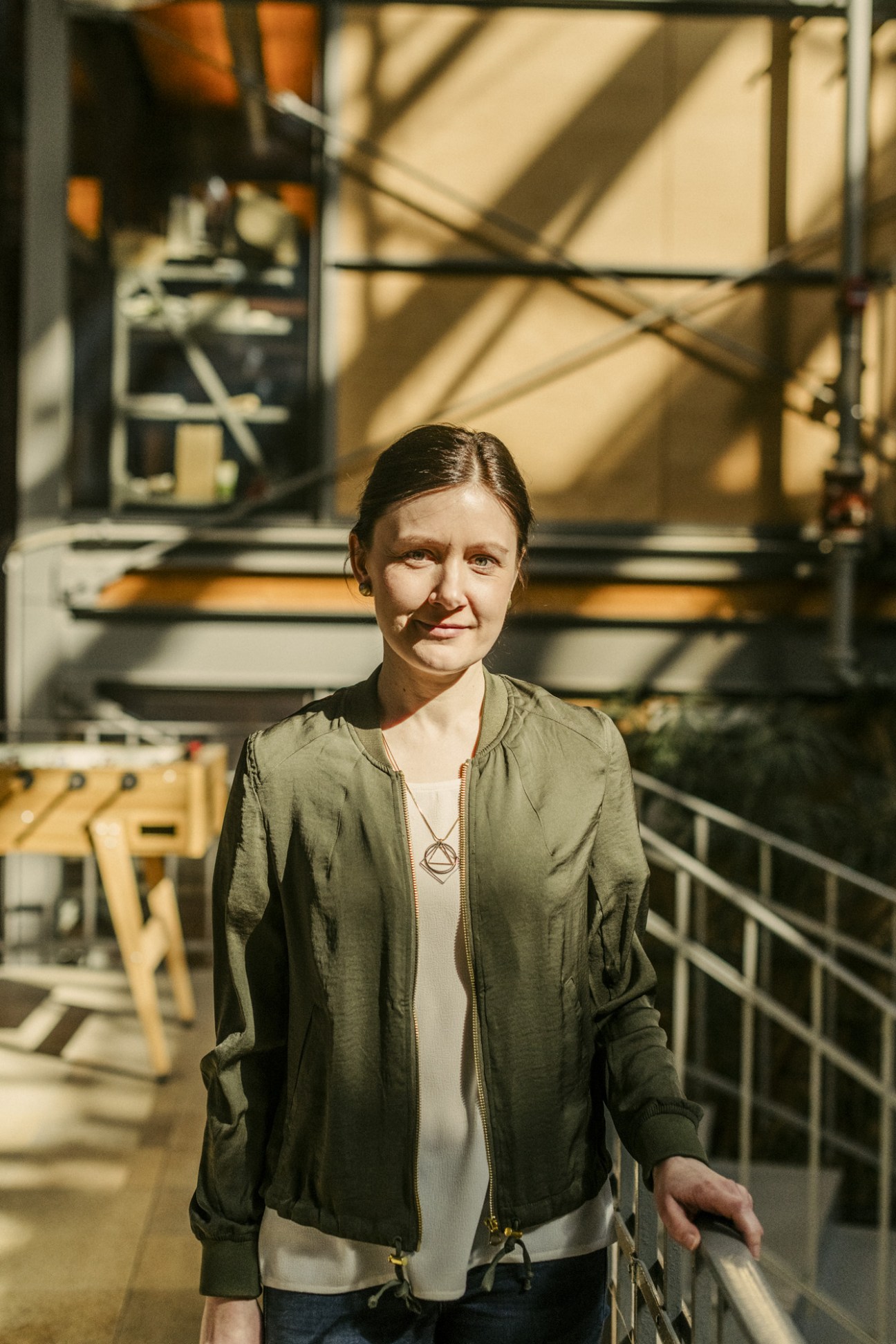 Woman wearing olive-green jacket standing in industrial building with geometric windows and scaffolding.