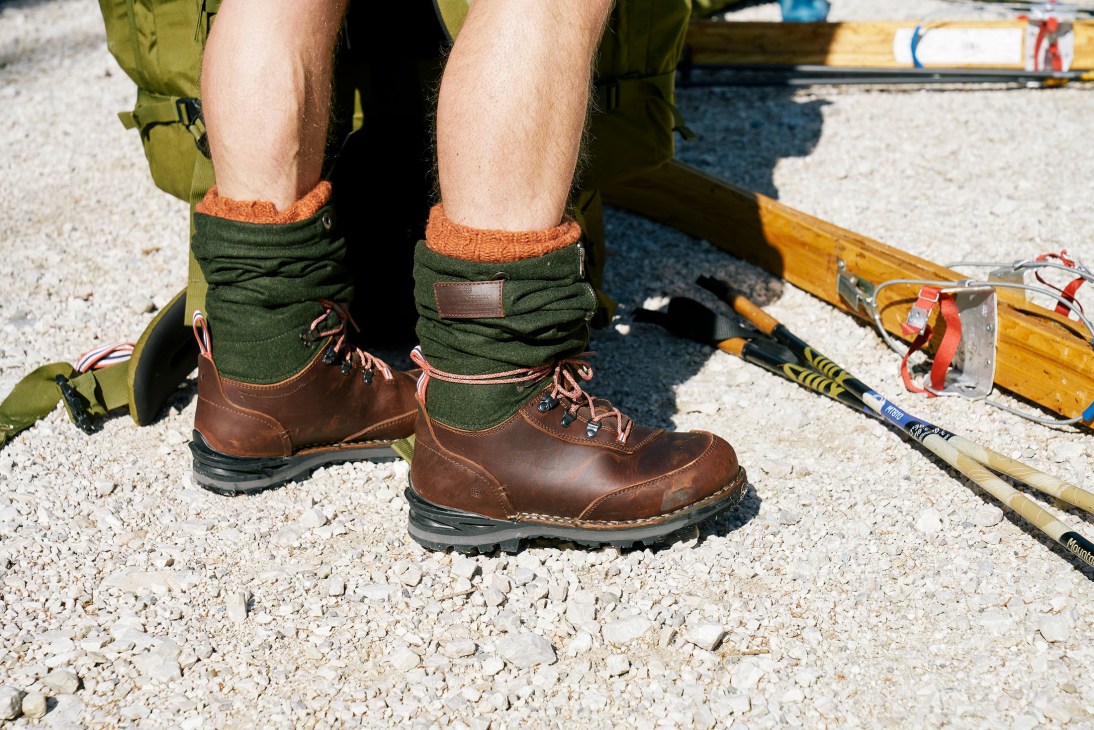 Brown leather hiking boots with green wool socks and orange cuff trim, worn while standing on gravel.