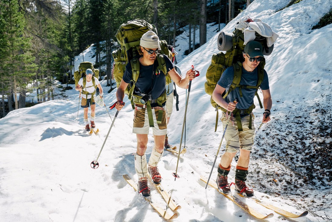 Three men in outdoor gear with large backpacks and ski poles hiking uphill through snow in a mountainous forest