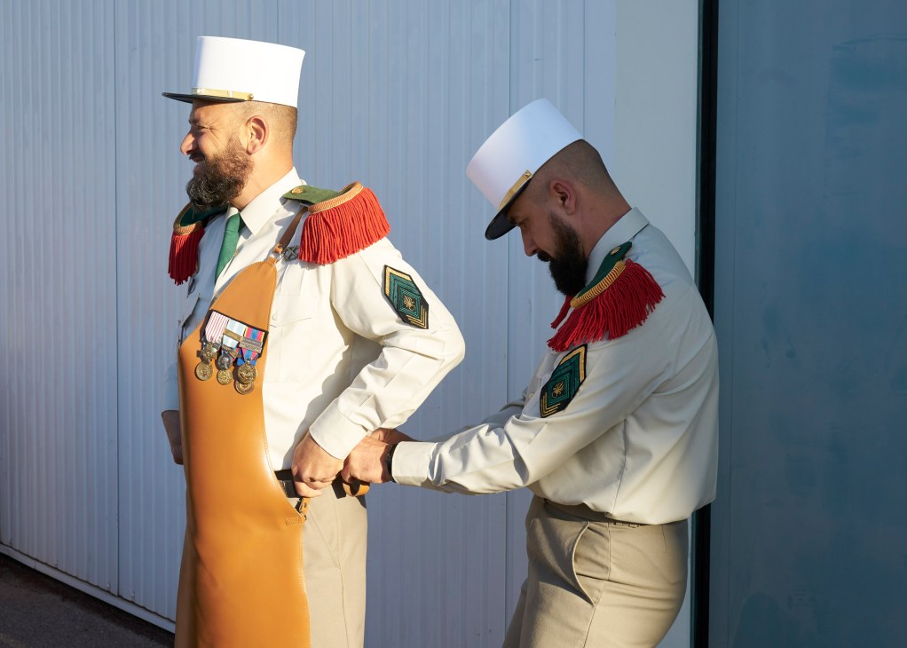 Two French Foreign Legion sappers in dress uniform adjusting tan leather aprons before a ceremonial parade.